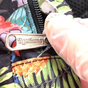 May include: Close-up of a black fabric with a colorful floral pattern. A silver zipper is partially open, revealing the zipper pull with the word "Loungefly" engraved on it. The fabric has red, green, blue, and purple flowers.