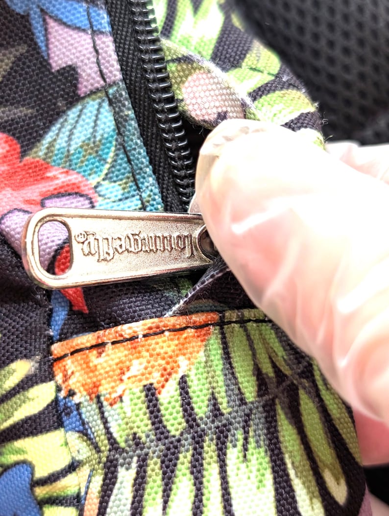 May include: Close-up of a black fabric with a colorful floral pattern. A silver zipper is partially open, revealing the zipper pull with the word "Loungefly" engraved on it. The fabric has red, green, blue, and purple flowers.