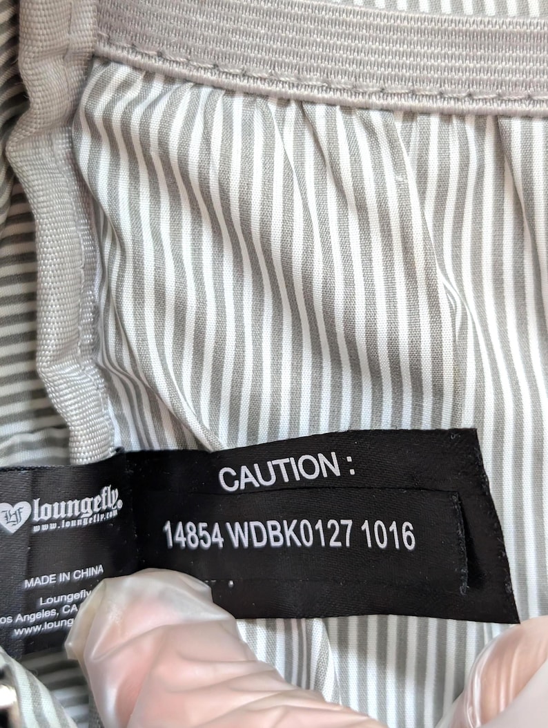 May include: Close-up of a gray and white striped fabric with a black tag. The tag reads "CAUTION:" followed by a product code. The brand name "Loungefly" is also visible on the tag. The fabric appears to be part of a bag or similar item.