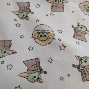 Op de afbeelding: Witte stof met een herhaald patroon van een groen buitenaards personage met grote oren en een bruine mantel, afgewisseld met kleine witte sterren. De alien wordt in verschillende poses afgebeeld.