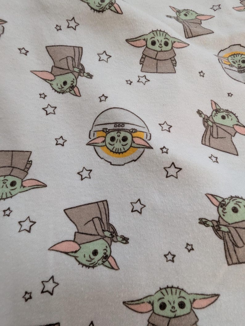 Op de afbeelding: Witte stof met een herhaald patroon van een groen buitenaards personage met grote oren en een bruine mantel, afgewisseld met kleine witte sterren. De alien wordt in verschillende poses afgebeeld.