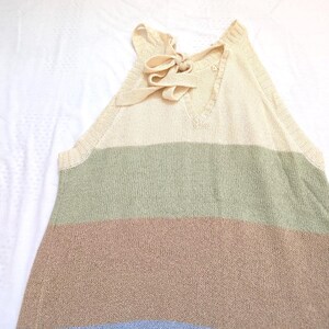 May include: A sleeveless, striped sweater in cream, green, brown, and blue. The top has a V-neck and a bow detail at the back of the neck. The sweater is made of a soft, knitted material.