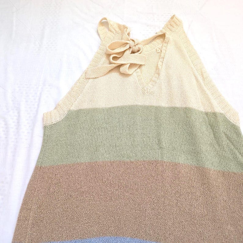 May include: A sleeveless, striped sweater in cream, green, brown, and blue. The top has a V-neck and a bow detail at the back of the neck. The sweater is made of a soft, knitted material.
