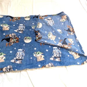 May include: A blue fleece blanket featuring characters from a popular science fiction franchise. The blanket has images of a character in armor, a green-skinned baby, and a robot. White stars are scattered across the blanket.
