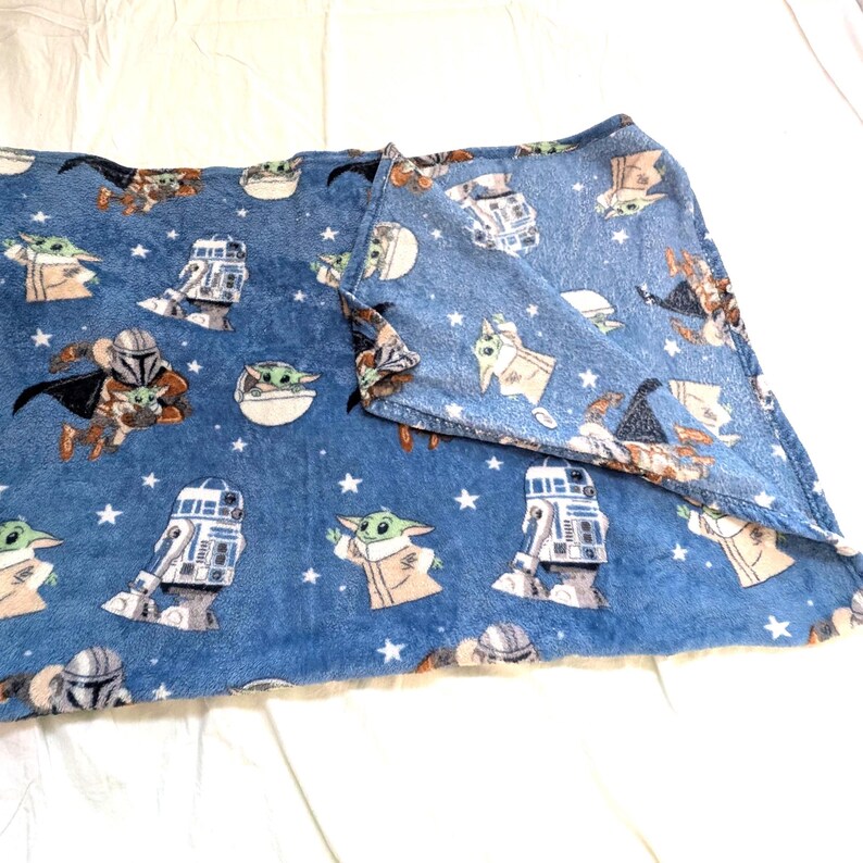 May include: A blue fleece blanket featuring characters from a popular science fiction franchise. The blanket has images of a character in armor, a green-skinned baby, and a robot. White stars are scattered across the blanket.