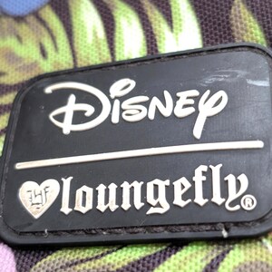 May include: Close-up of a black rectangular Disney Loungefly patch. The Disney logo is in white, above the Loungefly text. The background is a floral pattern with green and purple leaves.