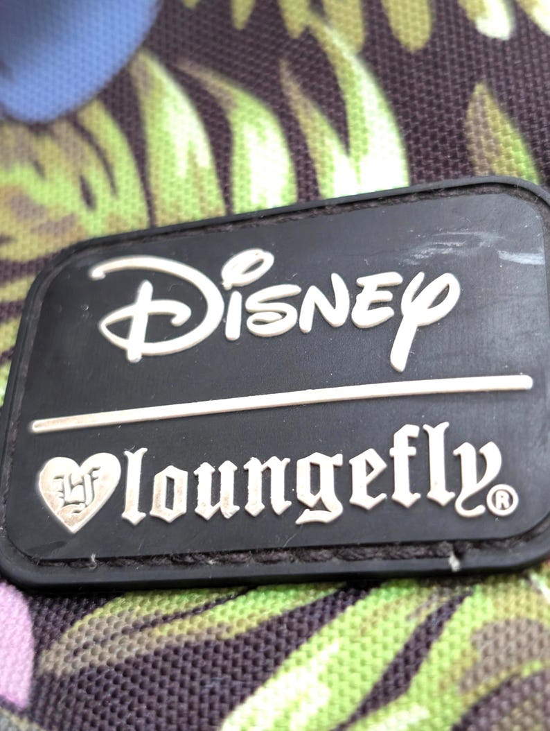 May include: Close-up of a black rectangular Disney Loungefly patch. The Disney logo is in white, above the Loungefly text. The background is a floral pattern with green and purple leaves.