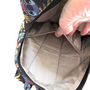 May include: Black backpack with a colorful Stitch character design. The interior features a gray and white striped lining. The backpack is open, revealing the inside. The zipper is black.
