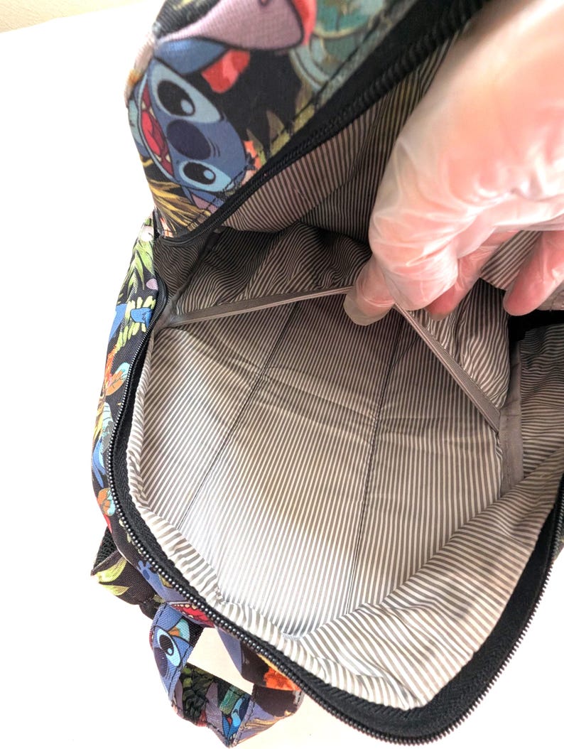 May include: Black backpack with a colorful Stitch character design. The interior features a gray and white striped lining. The backpack is open, revealing the inside. The zipper is black.