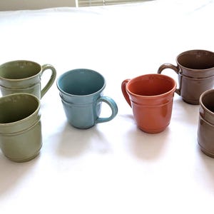 May include: Six ceramic mugs in various colors: green, blue, orange, and brown. Each mug has a handle and a simple, classic design. The mugs are arranged on a white surface, showcasing their different hues and shapes.