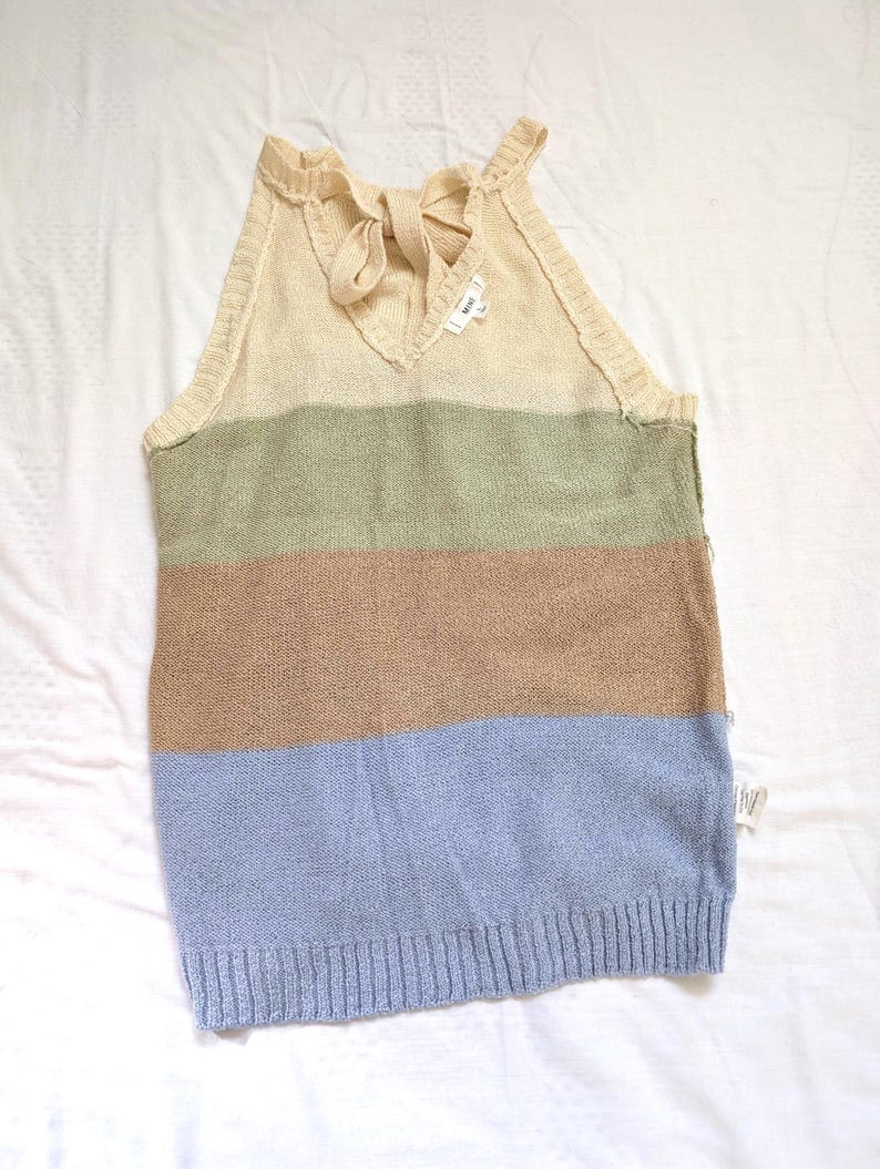 May include: A sleeveless, striped sweater in shades of beige, green, brown, and blue. The top features a V-neck with a bow detail and a ribbed hem. The sweater is made of a soft, knitted material.