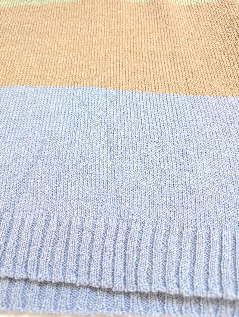 May include: Close-up of a knitted jumper with horizontal stripes. The jumper features bands of colour in light green, tan, and light blue. The knit fabric has a ribbed texture.