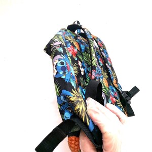 May include: A black backpack with a tropical print featuring blue cartoon characters, pineapples, and flowers. The backpack has black straps and a buckle. The word "Loungefly" is printed on the top of the backpack.