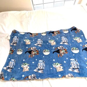May include: Blue blanket featuring Star Wars characters, including The Mandalorian, Grogu, and R2-D2, with white stars. The blanket is made of a soft material and is designed for comfort. The blanket is rectangular in shape.