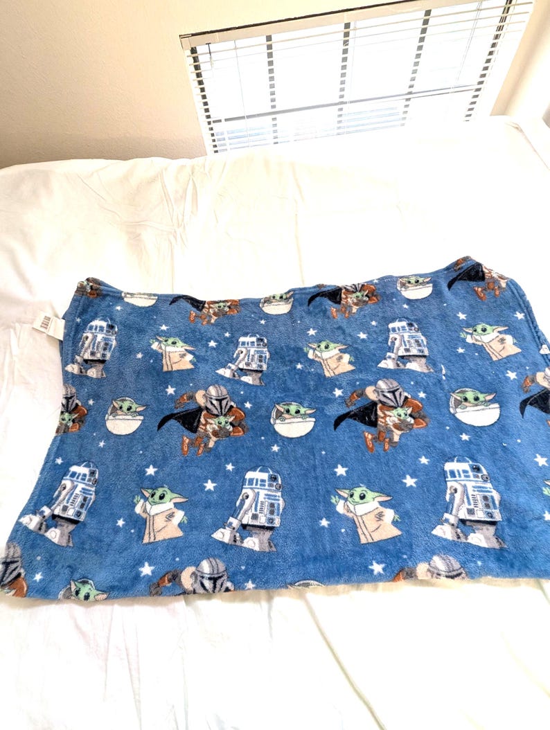 May include: Blue blanket featuring Star Wars characters, including The Mandalorian, Grogu, and R2-D2, with white stars. The blanket is made of a soft material and is designed for comfort. The blanket is rectangular in shape.