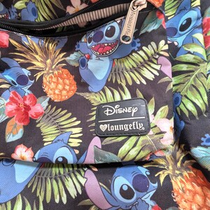 May include: A black backpack with a tropical pattern featuring the Disney character Stitch, pineapples, and flowers. The backpack has a zippered pocket and a Disney Loungefly label.