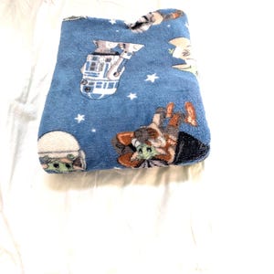 May include: A folded blue plush blanket featuring Star Wars characters and white stars. The blanket has images of R2-D2, Grogu, and other characters from the Star Wars franchise. The blanket is folded on a white surface.