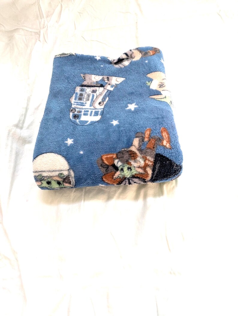 May include: A folded blue plush blanket featuring Star Wars characters and white stars. The blanket has images of R2-D2, Grogu, and other characters from the Star Wars franchise. The blanket is folded on a white surface.