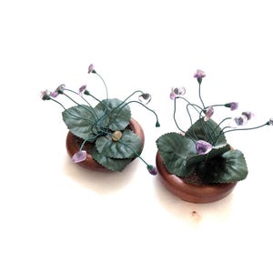 May include: Two small, decorative potted plants with dark green leaves and delicate stems bearing purple and clear crystal-like flowers. The plants are in round, wooden pots, set against a white background.