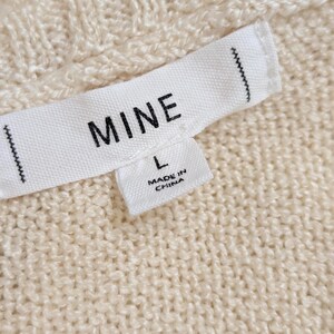 May include: Close-up of a cream-coloured knit fabric with a label that reads "MINE" and "L". The label also states "MADE IN CHINA". The fabric has a textured, cable-knit pattern.