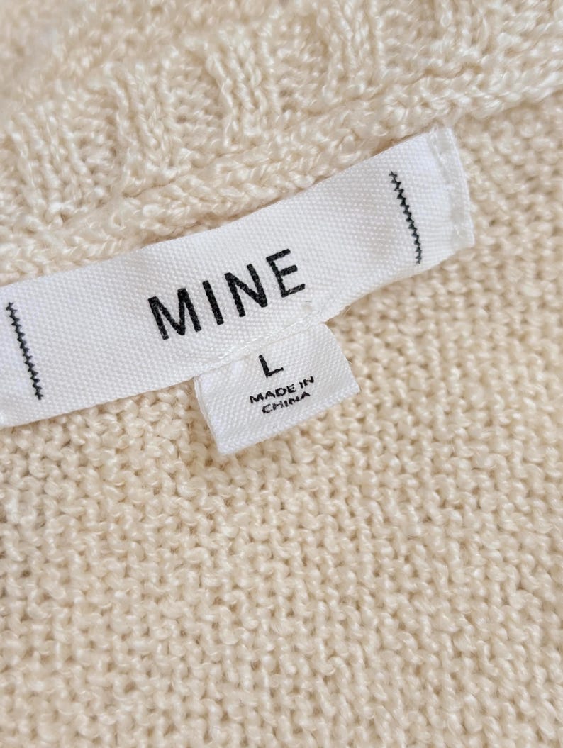 May include: Close-up of a cream-coloured knit fabric with a label that reads "MINE" and "L". The label also states "MADE IN CHINA". The fabric has a textured, cable-knit pattern.