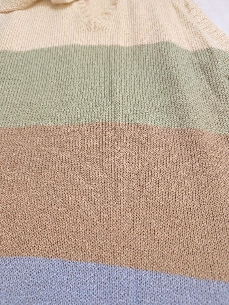 May include: Close-up of a knitted sweater with a V-neck collar. The sweater has horizontal stripes in cream, sage green, tan, and light blue. The knit fabric has a textured appearance.