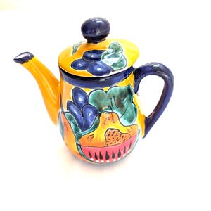 May include: A hand-painted ceramic teapot with a yellow body, a blue handle, and a lid. The teapot is decorated with colorful floral and abstract designs in shades of blue, green, and red. The spout is yellow.