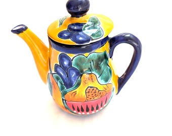 May include: A hand-painted ceramic teapot with a yellow body, a blue handle, and a lid. The teapot is decorated with colorful floral and abstract designs in shades of blue, green, and red. The spout is yellow.