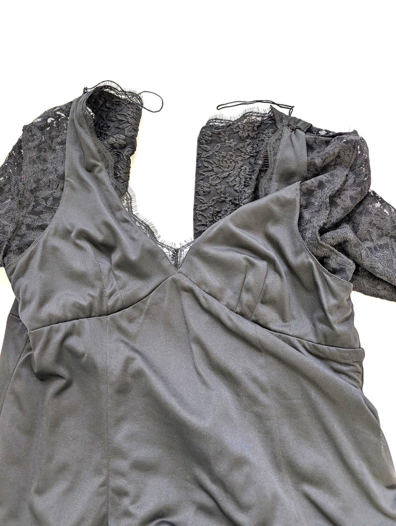 May include: A black satin dress with lace sleeves. The dress features a V-neckline and a fitted bodice. The sleeves are made of a delicate black lace. The dress is laid flat on a white surface.