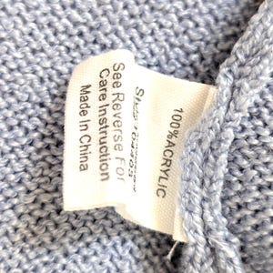 May include: Close-up of a pale blue knitted textile with a white label. The label states "100% ACRYLIC" and "See Reverse For Care Instruction Made In China."