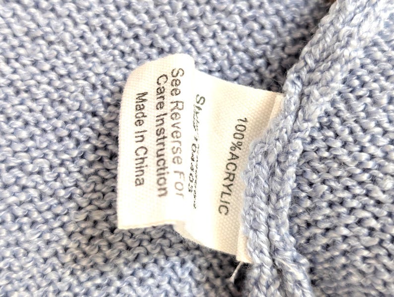 May include: Close-up of a pale blue knitted textile with a white label. The label states "100% ACRYLIC" and "See Reverse For Care Instruction Made In China."