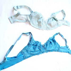 May include: Two light blue bras are displayed against a white background. The top bra is a pale blue with lace detailing, while the bottom bra is a brighter blue with intricate embroidery. Both bras have adjustable straps and underwire support.