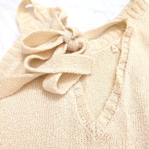 May include: A close-up shot of a cream-coloured knitted top featuring a V-neck and a bow detail at the neckline. The knitwear has a textured appearance and a subtle green stripe at the bottom. The bow is tied in the centre, adding a decorative element to the garment.