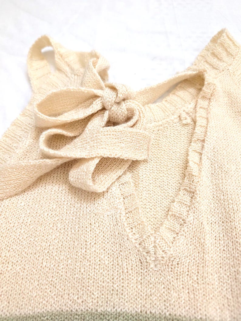 May include: A close-up shot of a cream-coloured knitted top featuring a V-neck and a bow detail at the neckline. The knitwear has a textured appearance and a subtle green stripe at the bottom. The bow is tied in the centre, adding a decorative element to the garment.