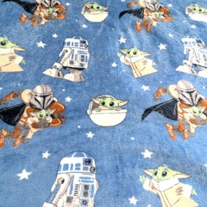 May include: A blue plush blanket featuring repeating patterns of characters from a popular science fiction franchise. The designs include a character in a Mandalorian helmet, a green alien, and a droid, all set against a background of white stars.