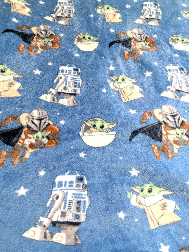 May include: A blue plush blanket featuring repeating patterns of characters from a popular science fiction franchise. The designs include a character in a Mandalorian helmet, a green alien, and a droid, all set against a background of white stars.
