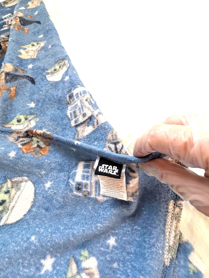May include: Blue Star Wars fleece blanket featuring Grogu and other characters, with a white star pattern. The blanket has a tag with the Star Wars logo.