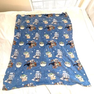 May include: Blue blanket featuring a repeating pattern of characters from a popular science fiction series. The design includes images of a Mandalorian, a small green alien, and a droid, along with white stars. The blanket is made of a soft, plush material.