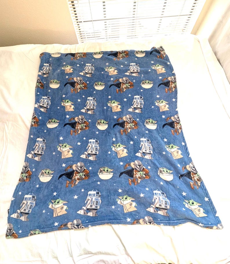 May include: Blue blanket featuring a repeating pattern of characters from a popular science fiction series. The design includes images of a Mandalorian, a small green alien, and a droid, along with white stars. The blanket is made of a soft, plush material.