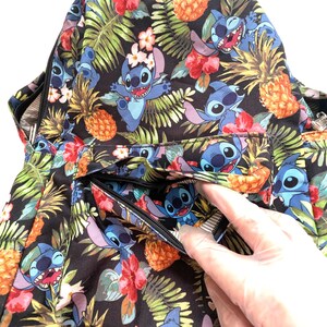 May include: A black backpack with a tropical pattern featuring blue cartoon characters, pineapples, and flowers. The backpack has a zippered pocket and a black zipper. The design includes green leaves and red and pink flowers.