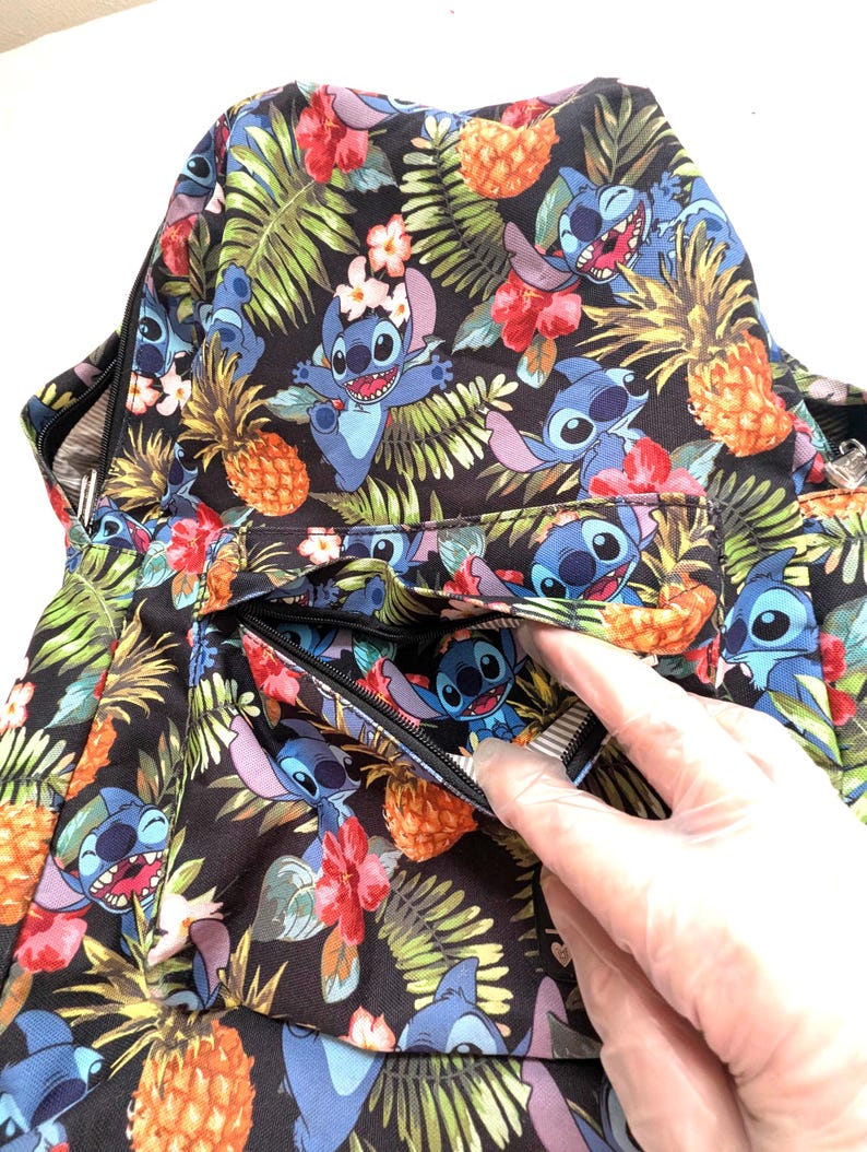 May include: A black backpack with a tropical pattern featuring blue cartoon characters, pineapples, and flowers. The backpack has a zippered pocket and a black zipper. The design includes green leaves and red and pink flowers.