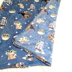 May include: Blue plush blanket featuring a repeating pattern of Star Wars characters, including Grogu, R2-D2, and Mandalorian figures. The blanket has a soft texture and is decorated with white stars.