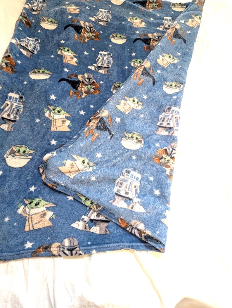 May include: Blue plush blanket featuring a repeating pattern of Star Wars characters, including Grogu, R2-D2, and Mandalorian figures. The blanket has a soft texture and is decorated with white stars.