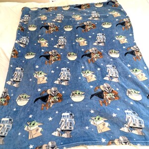 May include: Blue plush blanket featuring repeating patterns of The Mandalorian, Grogu, and R2-D2 characters, along with white stars. The blanket is soft and cozy, perfect for fans of the Star Wars series.