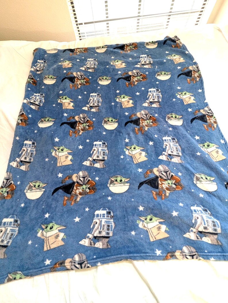 May include: Blue plush blanket featuring repeating patterns of The Mandalorian, Grogu, and R2-D2 characters, along with white stars. The blanket is soft and cozy, perfect for fans of the Star Wars series.