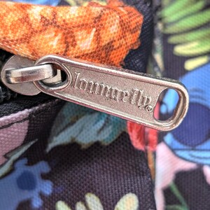 May include: Close-up of a black bag with a colorful floral and character print. A silver zipper pull with the brand name "Loungefly" is visible. The bag features a vibrant design with shades of blue, orange, green, and pink.