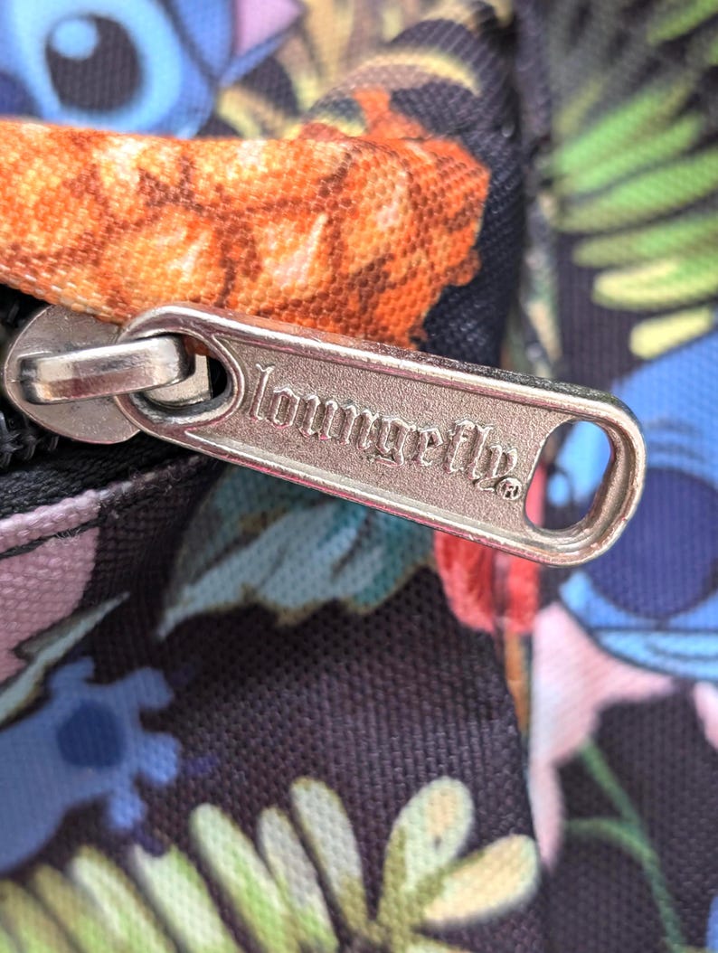 May include: Close-up of a black bag with a colorful floral and character print. A silver zipper pull with the brand name "Loungefly" is visible. The bag features a vibrant design with shades of blue, orange, green, and pink.