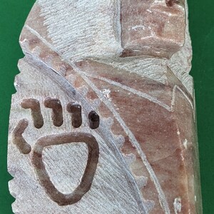 May include: A carved stone sculpture of a person's head with a bear paw print on the side. The sculpture is made of a light brown stone with a reddish hue.