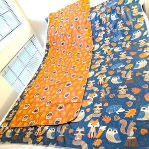 May include: A reversible blanket featuring an orange side with owl patterns and a blue side with woodland animal designs, including foxes, raccoons, and squirrels. The blanket is made of a soft material and is suitable for a child's room.