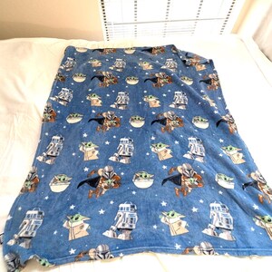 May include: A blue blanket featuring a repeating pattern of characters from a popular science fiction franchise. The design includes images of a bounty hunter, a small green alien, and a droid, along with white stars.
